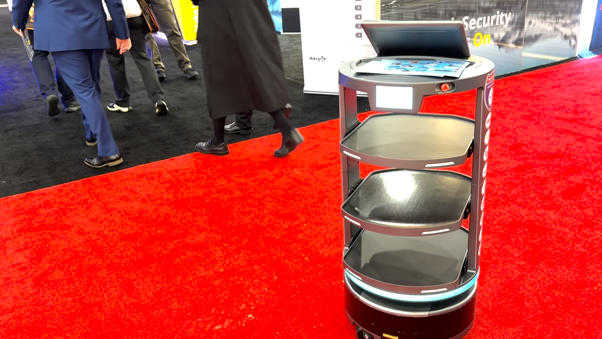 Autonomous service robot with three shelves on a red carpet at a trade show, laptop on top and people walking in the background