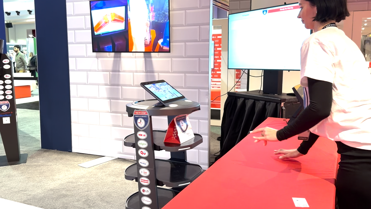Trade show booth with a touchscreen tablet on a tiered pedestal and a large monitor on a white brick wall; a woman in white interacts at a red counter.