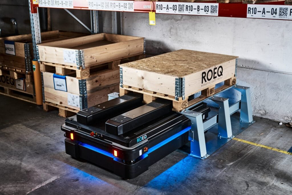 Autonomous pallet robot (ROEQ) under a wooden crate on a metal rack in a warehouse, blue underglow.