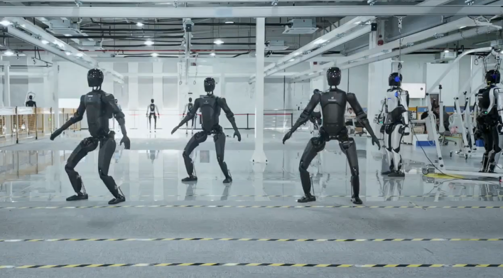 Four black humanoid robots practicing synchronized movement in a bright, industrial-lab gym with a glossy floor and safety tape on the ground.