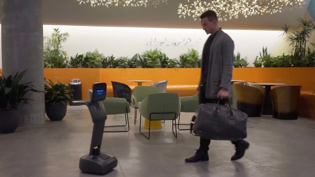 Man in a gray coat walks with a large black bag through a modern, plant-filled lounge area with orange seating behind him