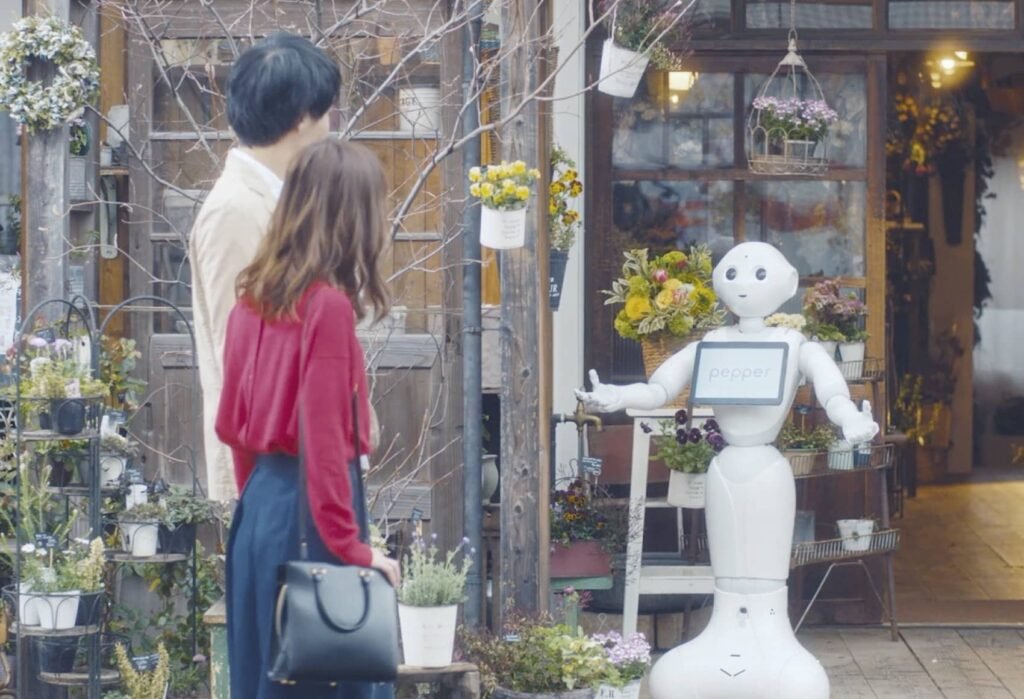 Two people with backs to the camera stand outside a flower shop as a white Pepper robot greets customers near the entrance, a display of plants all around.