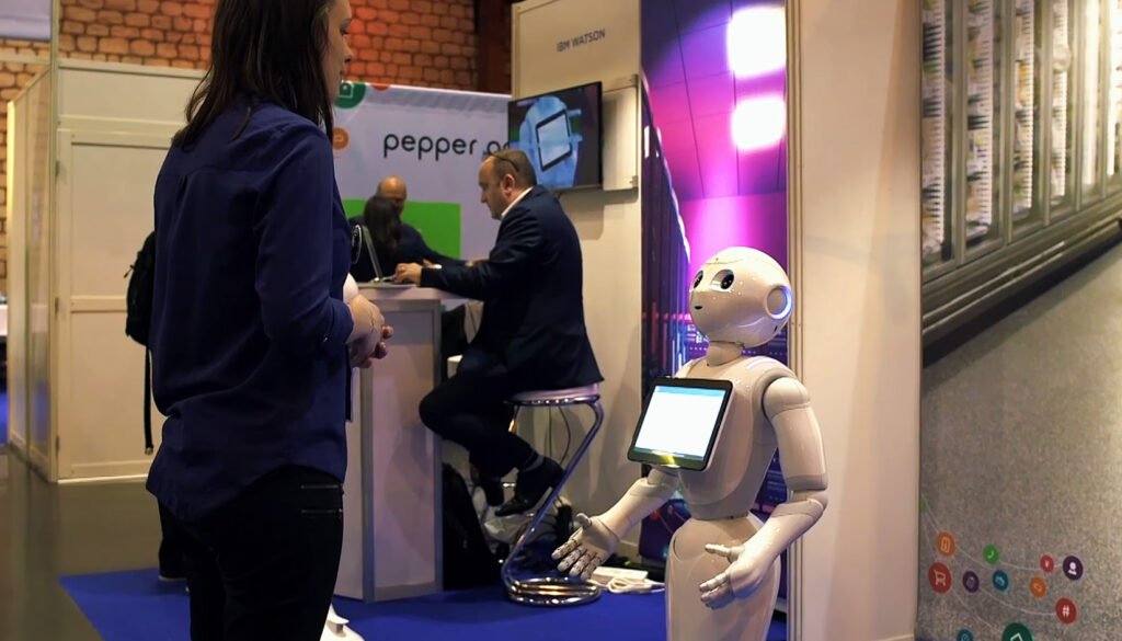Woman in a blue blouse stands at a booth, talking to a white humanoid robot with a touchscreen tablet on its chest.