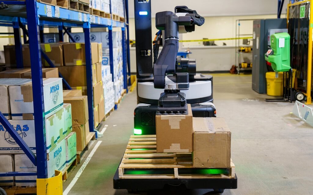 Automated warehouse robot arm placing a box onto a pallet in a stocked aisle with shelves and yellow safety tape nearby.