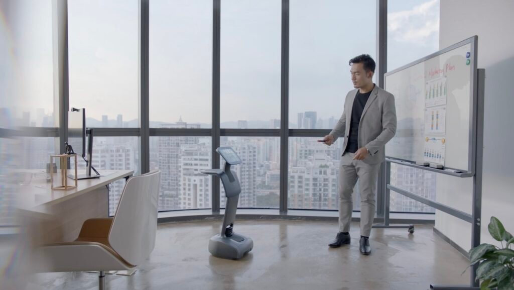 Presenter in a gray suit uses a remote next to a mobile robot in a glass-walled office, with charts on a whiteboard behind him.