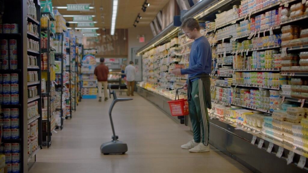Man in blue shirt and green pants holds a red basket while inspecting a floor-cleaning robot in a grocery store aisle.