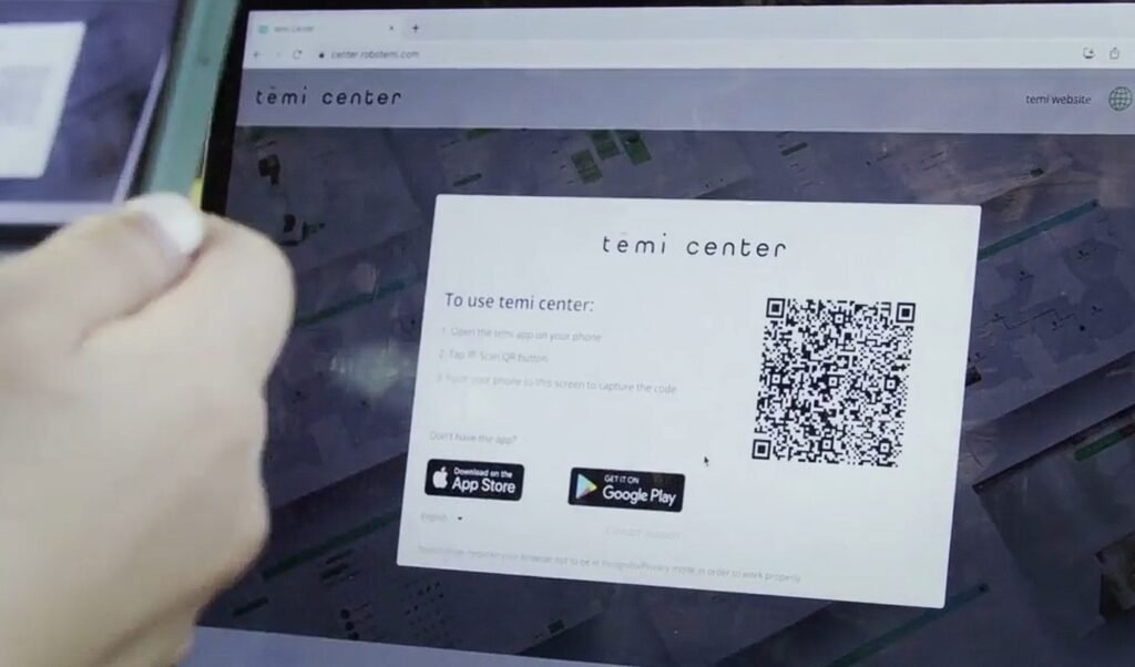 Close-up of a laptop screen showing the 'temi center' page; a card with a large QR code and App Store/Google Play badges is displayed, with a hand nearby
