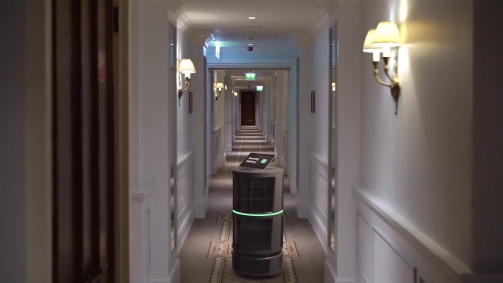 Autonomous delivery robot standing in a hotel hallway with doors on both sides and wall lamps."] ,