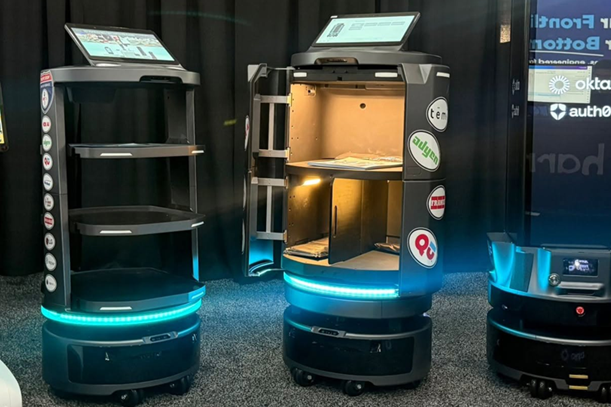 Three autonomous delivery robots with circular blue LED bases and touchscreen panels on top, on display in a tech showroom.