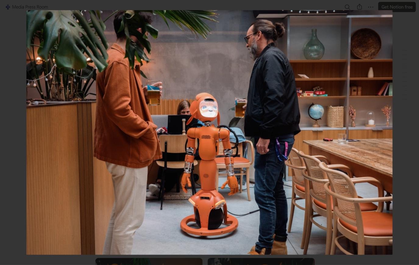 Orange humanoid robot stands between two adults in a cozy cafe-like room.